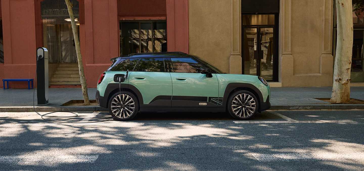 Zijaanzicht van de MINI Aceman Electric in Ocean Wave Green met dak in Black, die op straat wordt opgeladen.