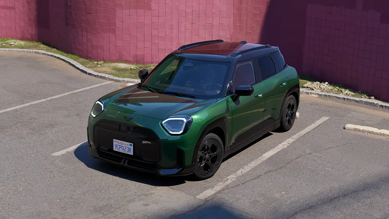 Vooraanzicht van een MINI Aceman Electric in British Racing Green op een parkeerplaats met een roze muur op de achtergrond