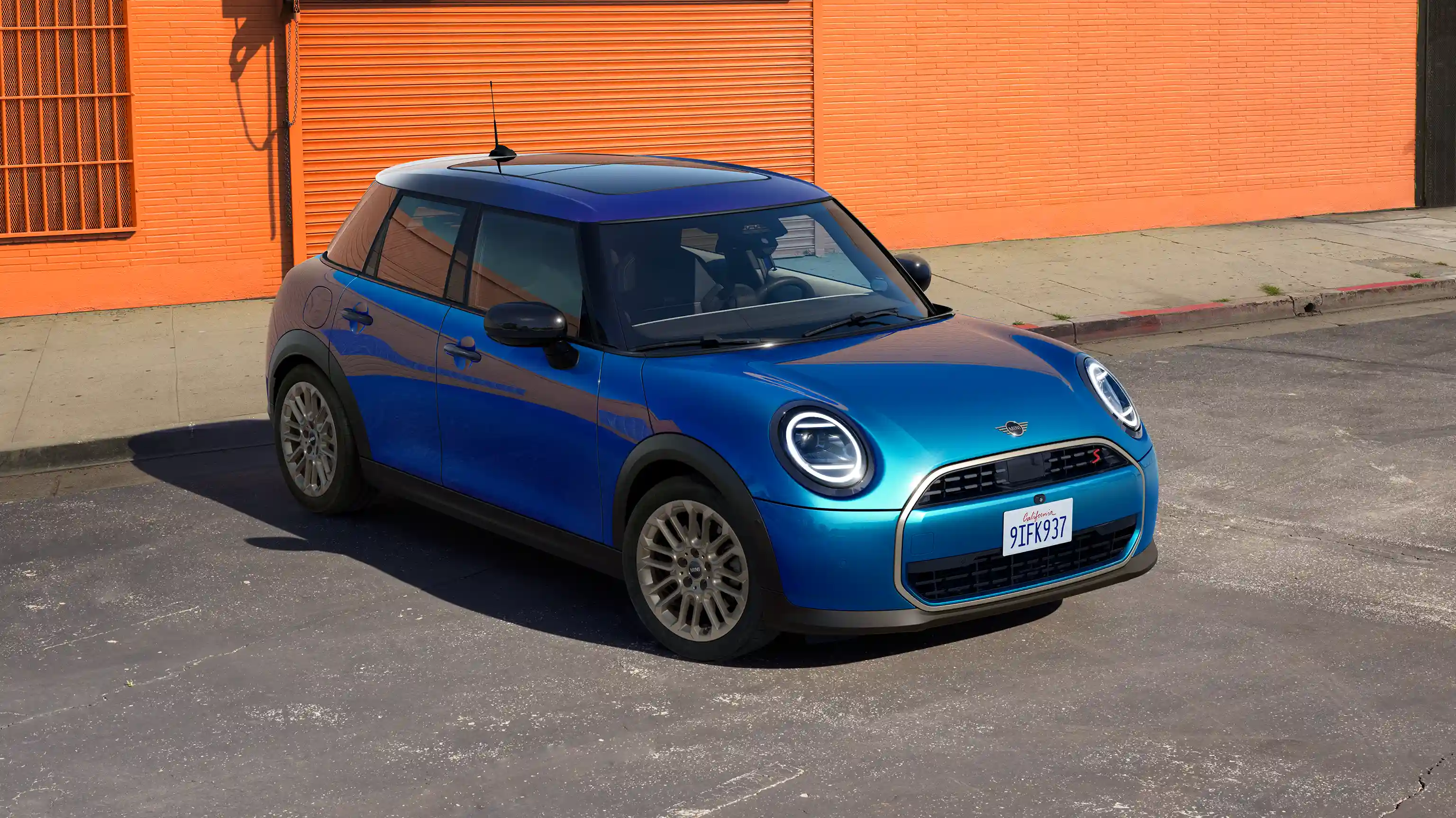Vooraanzicht van parkeren van MINI Cooper 5-Deurs in Icy Sunshine Blue met dak in Multitone Blue op straat met een oranje muur op de achtergrond