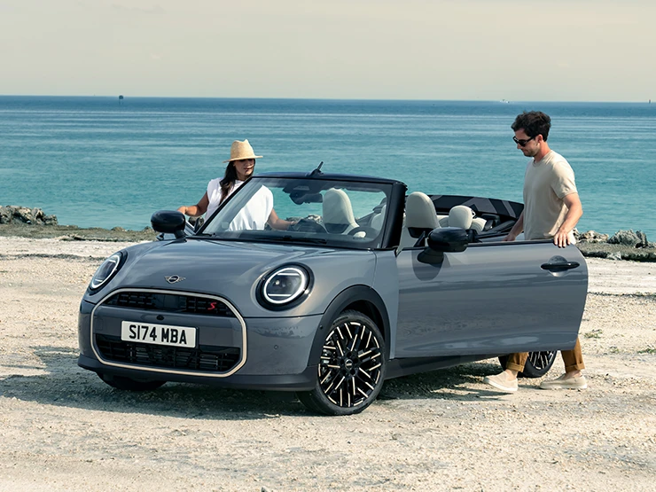Aanzicht links van voren op de MINI Cooper Cabrio in Copper Grey op een strand met twee personen ernaast.