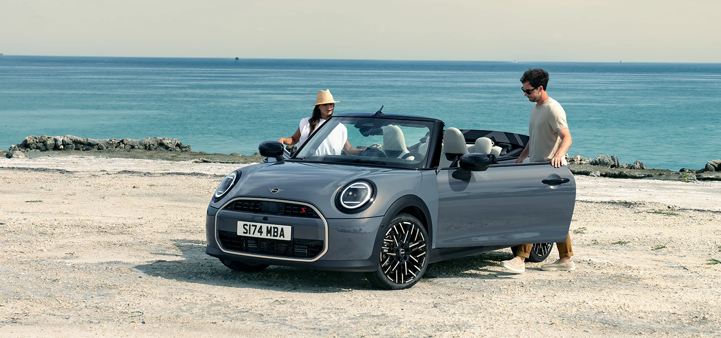 Aanzicht links van voren op de MINI Cooper Cabrio in Copper Grey op een strand met twee personen ernaast.