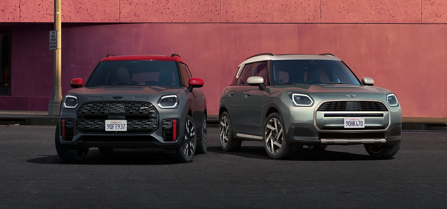 Vooraanzicht van de MINI Countryman JCW in Legend Grey met dak in Red en de MINI Countryman in Smokey Green met dak in Vibrant Silver naast elkaar.