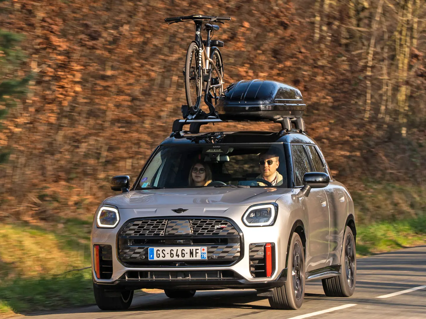 MINI Countryman Cooper in Silver op een bergweg met MINI dakdrager, MINI dakkoffer, MINI dakfietsdrager met daarop één fiets en MINI John Cooper Works winglet – vooraanzicht. 