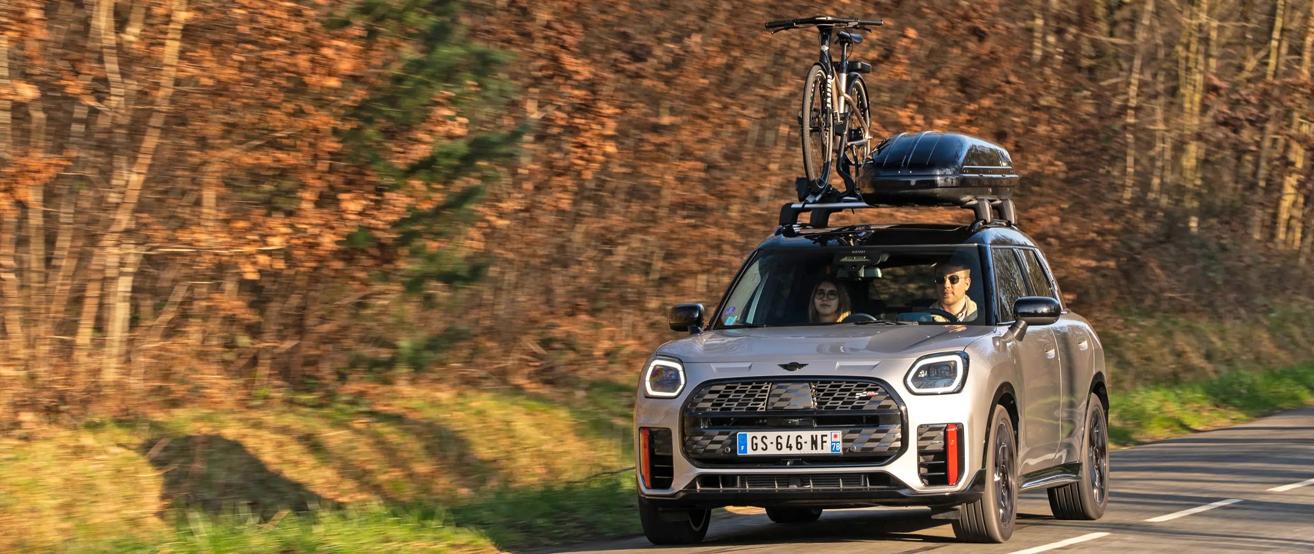 MINI Countryman Cooper in Silver op een bergweg met MINI dakdrager, MINI dakkoffer, MINI dakfietsdrager met daarop één fiets en MINI John Cooper Works winglet – vooraanzicht.