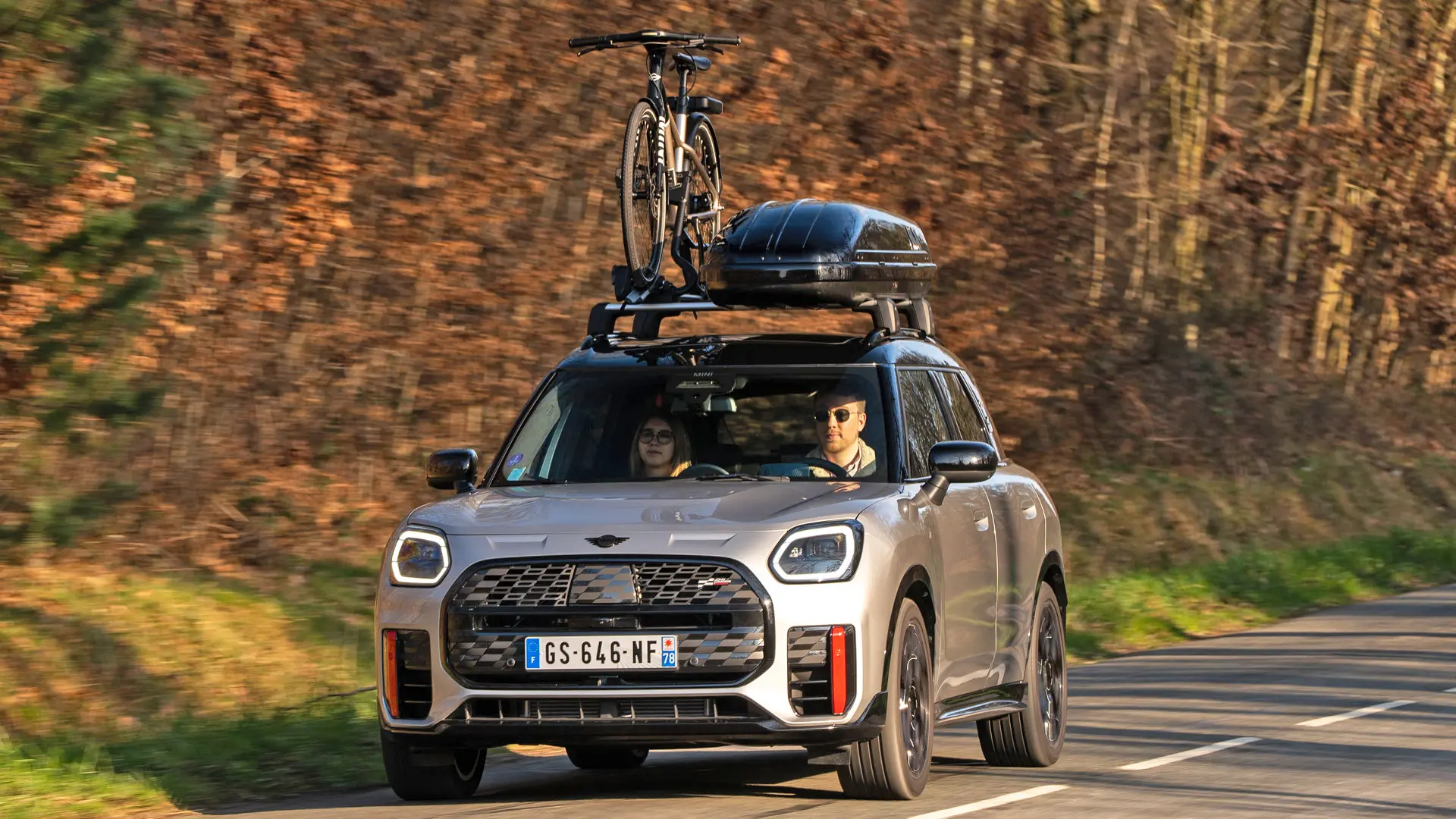 MINI Countryman Cooper in Silver op een bergweg met MINI dakdrager, MINI dakkoffer, MINI dakfietsdrager met daarop één fiets en MINI John Cooper Works winglet – vooraanzicht.