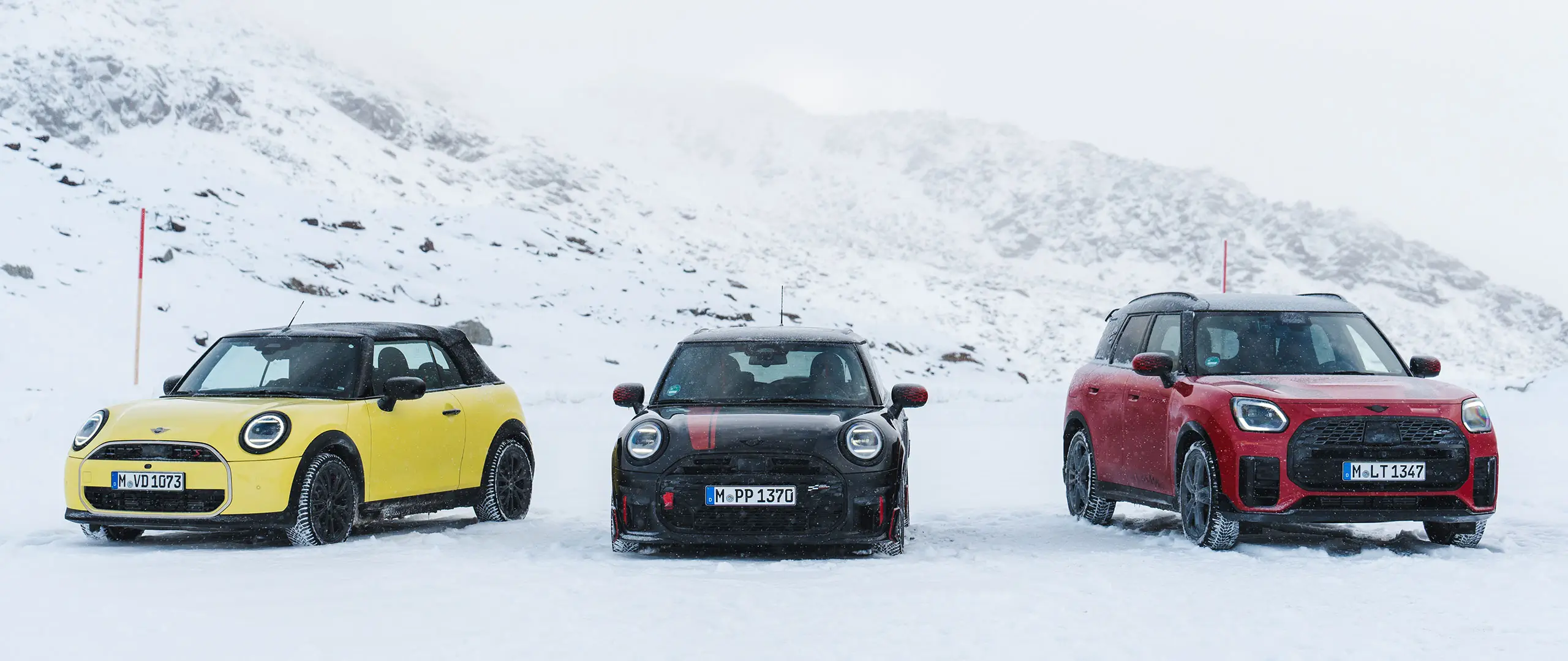 MINI Cooper Cabrio in Sunny Side Yellow, MINI John Cooper Works in Midnight Black II en MINI Countryman Cooper in Chili Red — stilstaand op een besneeuwde bergweg, vooraanzicht.