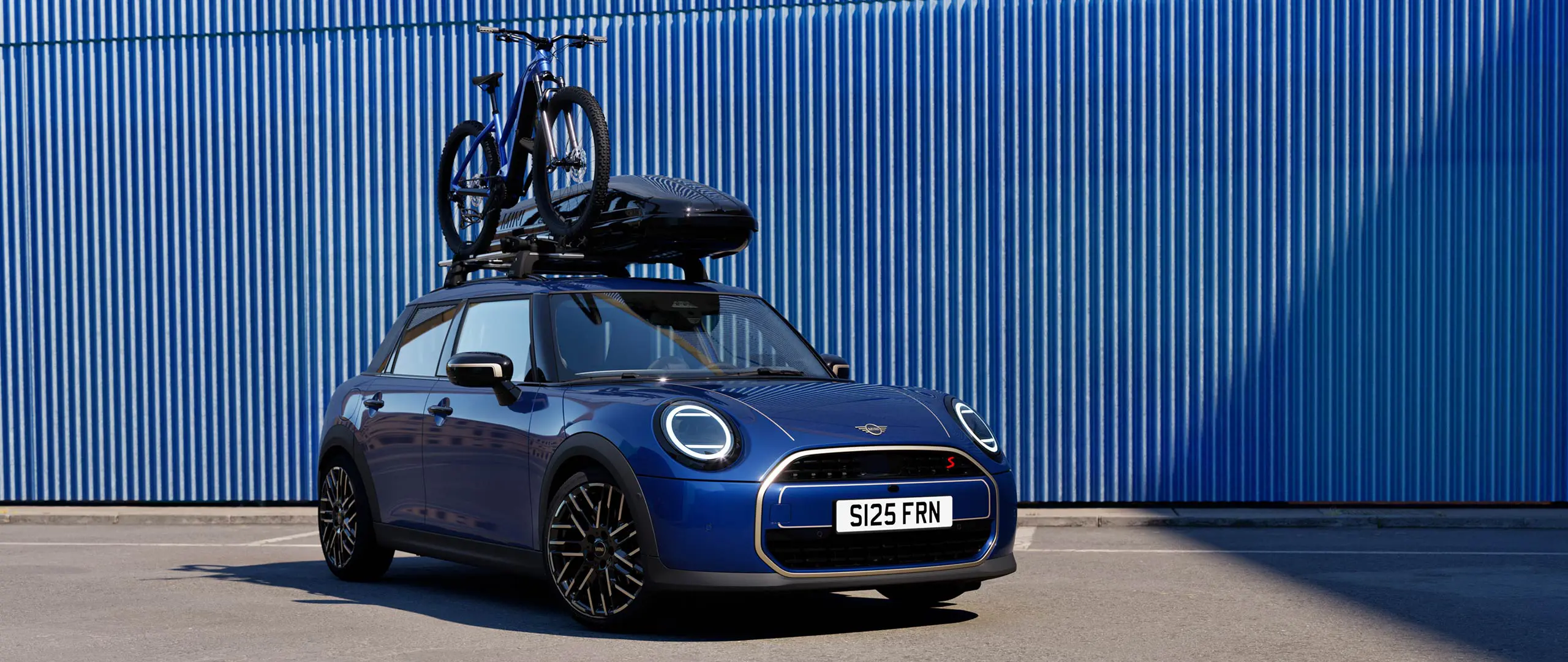 Vooraanzicht van MINI Aceman in Indigo Sunset Blue met MINI dakfietsendrager met mountainbike en MINI dakkoffer 320.
