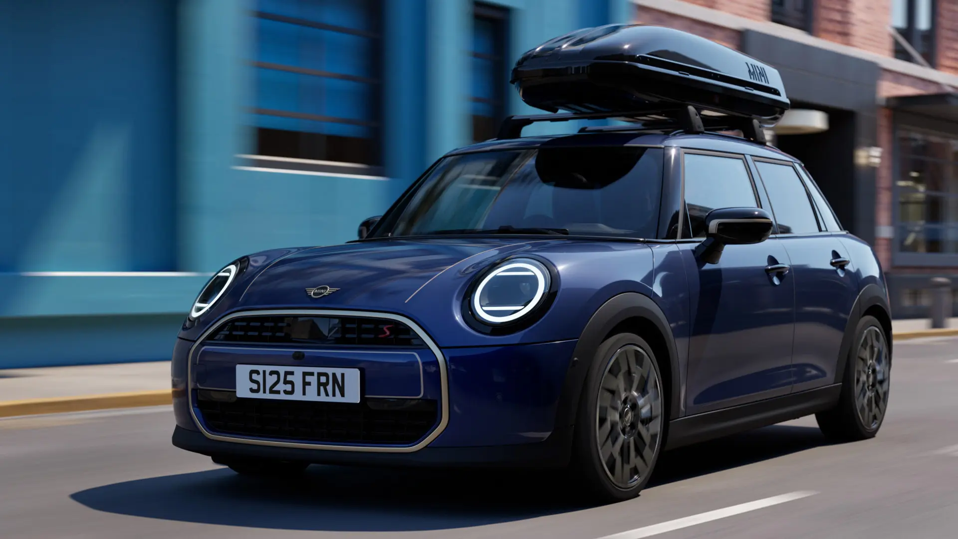 Driekwart vooraanzicht van MINI Aceman in Indigo Sunset Blue, uitgerust met MINI dakdragers met een MINI dakkoffer 320. 