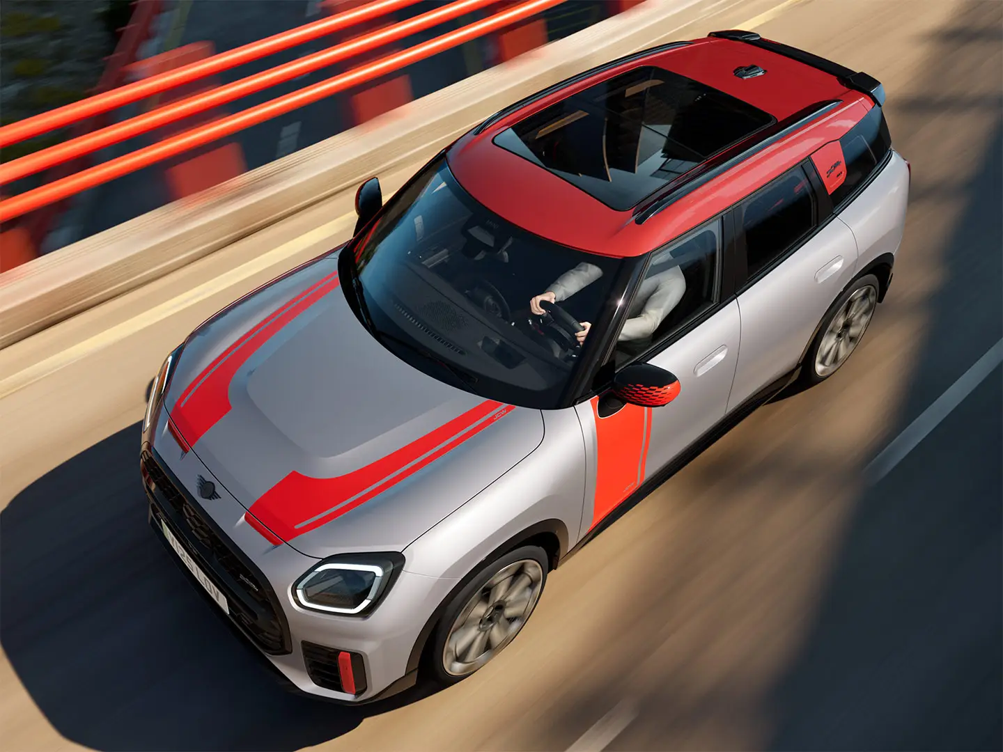 MINI Countryman John Cooper Works in Melting Silver met buitenspiegelkap, zijfoliëring, motorkapstickers, dak en winglet in Chili Red, rijdt in de stad — zijaanzicht van voor.