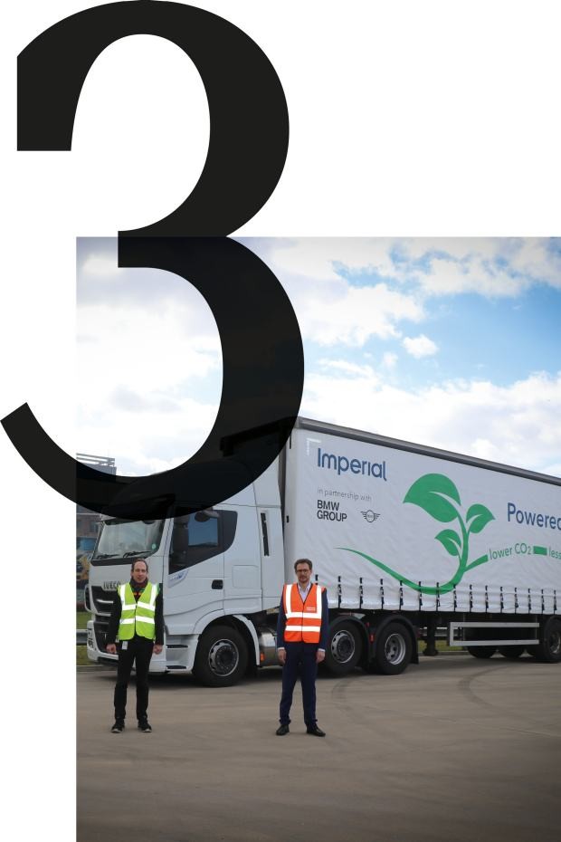 Foto van een vrachtwagen voor de MINI-fabriek in Oxford, die in het kader van de duurzame vervoerslogistiek met vloeibaar aardgas (LNG) wordt aangedreven en waarvoor drie werknemers staan die een hesje met hoge zichtbaarheid dragen. 